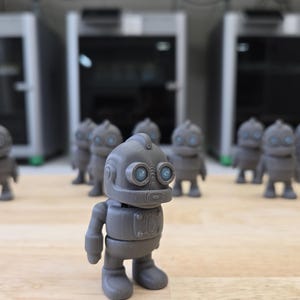 May include: A collection of gray robot figurines with blue eyes, standing on a wooden surface. The robots have a retro design, with rounded bodies and large feet. Several 3D printers are in the background.