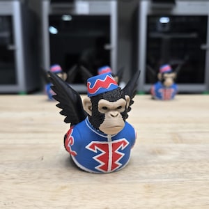 May include: A collection of whimsical monkey figurines. Each figurine is dressed in a blue jacket with red accents and a matching hat. The monkeys have black wings and are arranged on a wooden surface.