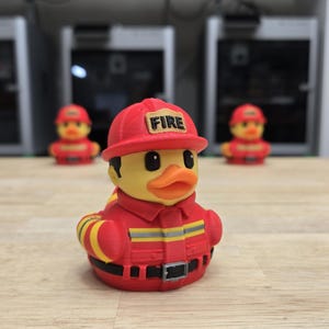 May include: A yellow rubber duck wearing a red firefighter uniform and helmet with the word "FIRE". The duck has black eyes and an orange beak. Other firefighter ducks are in the background.