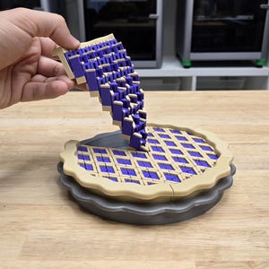 May include: A 3D-printed pie model with a lattice crust design. The pie filling is a deep purple color, and the crust is tan. A hand holds a section of the pie, showing the filling cascading into the pie.