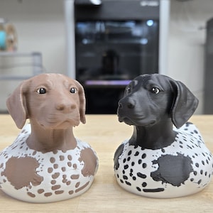 May include: Two dog figurines, one brown and one black, each with a white base speckled with brown or black spots. The dogs have detailed features and are set on a wooden surface.