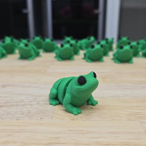 May include: A group of green frog figurines with black eyes. One frog is in the foreground, facing the viewer, while the rest are lined up in the background.