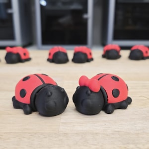 May include: A collection of red and black ladybug figurines. The ladybugs have a red shell with black spots and a black head. One ladybug has a red bow. The figurines are on a light brown surface.