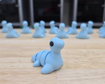 May include: A light blue, seal-like figurine with a black eye, lying on a wooden surface. Several similar figurines are in the background. The figurines appear to be handmade or 3D-printed.