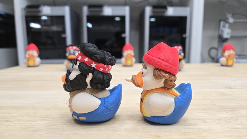 可能包含:两只新奇的鸭子模型。一只鸭子戴着红色头巾,头发乌黑,另一只鸭子戴着红色帽子,身穿橙色背心。两只鸭子均为蓝白相间,喙为橙色。背景中还有其他类似的鸭子模型。