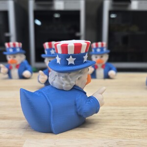 May include: A group of Uncle Sam-themed rubber ducks. Each duck is wearing a blue suit, a red and white striped top hat with stars, and a white beard. The ducks are displayed on a wooden surface.