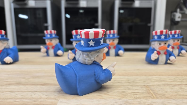 May include: A group of Uncle Sam-themed rubber ducks. Each duck is wearing a blue suit, a red and white striped top hat with stars, and a white beard. The ducks are displayed on a wooden surface.