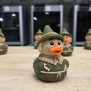 May include: A collection of novelty duck figurines, each dressed as a scarecrow. The ducks have orange beaks, black eyes, and wear green hats and outfits with tan accents. They are arranged on a light-colored wooden surface.