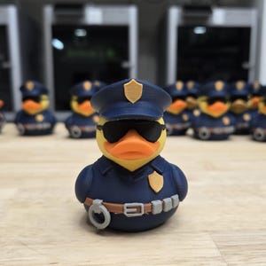 May include: A yellow rubber duck wearing a navy blue police uniform, sunglasses, and a hat. The duck has an orange beak and is surrounded by other ducks in similar uniforms. The ducks are on a light brown surface.