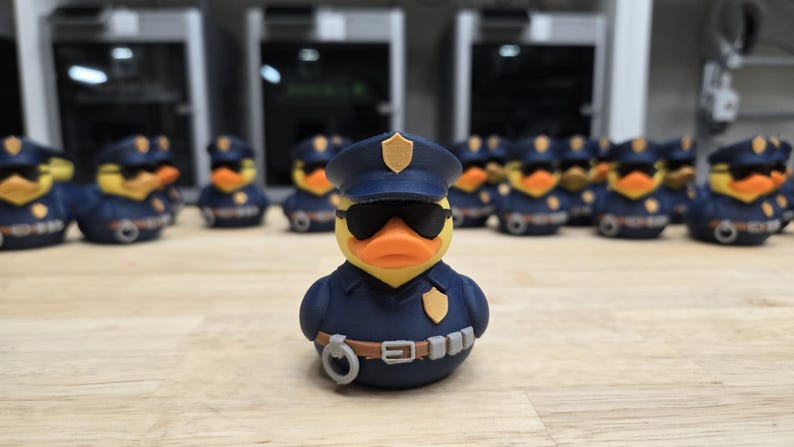 May include: A yellow rubber duck wearing a navy blue police uniform, sunglasses, and a hat. The duck has an orange beak and is surrounded by other ducks in similar uniforms. The ducks are on a light brown surface.