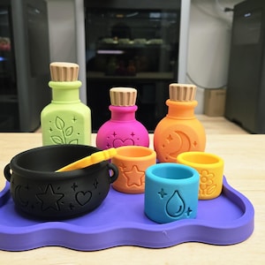 May include: A set of colorful potion-themed items including a black cauldron with a yellow spoon, a light green bottle, a pink bottle, an orange bottle, and three small cups in orange, yellow, and blue, all arranged on a purple wavy tray.