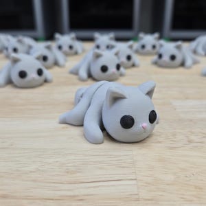 May include: A collection of gray cat figurines with black eyes and pink noses, lying on a light brown wooden surface. The figurines are in a relaxed pose, with their legs extended. The background features more of the same cat figurines.