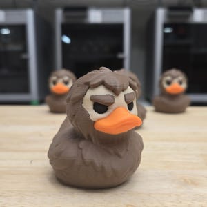 May include: A collection of brown rubber duck figurines with orange beaks and white eyes. The ducks have a grumpy expression. The ducks are arranged on a light brown wooden surface.