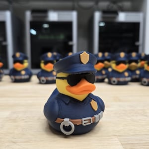 May include: A group of yellow rubber duckies dressed as police officers, wearing blue uniforms, hats, and sunglasses. The duck in the foreground is in focus, with others blurred in the background. The ducks have orange beaks and are on a wooden surface.