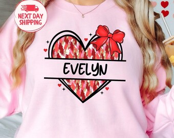 May include: Pink sweatshirt with a red and white heart design and the name "Evelyn" printed on it. The heart has a red bow on top.