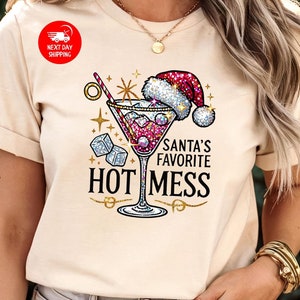 May include: A beige t-shirt with a glittery design of a martini glass with a Santa hat on top. The text "Santa's Favorite Hot Mess" is printed below the glass.