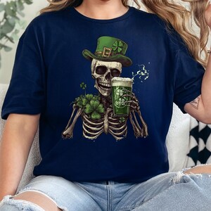 Puede incluir: Una camiseta azul marino con un esqueleto vestido de duende, con un sombrero verde y sosteniendo una jarra de cerveza verde. El esqueleto est&aacute; rodeado de tr&eacute;boles.
