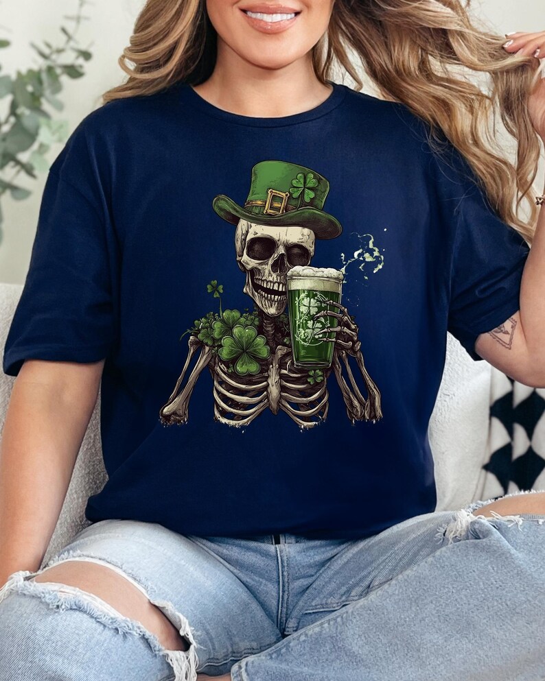 Puede incluir: Una camiseta azul marino con un esqueleto vestido de duende, con un sombrero verde y sosteniendo una jarra de cerveza verde. El esqueleto est&aacute; rodeado de tr&eacute;boles.