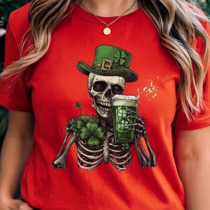 Puede incluir: Camiseta roja con un esqueleto vestido de duende verde, sosteniendo una jarra de cerveza verde. El esqueleto est&aacute; rodeado de tr&eacute;boles.