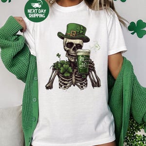 Puede incluir: Camiseta blanca con un esqueleto verde que lleva un sombrero de duende verde y sostiene una jarra de cerveza verde. El esqueleto est&aacute; rodeado de tr&eacute;boles verdes.