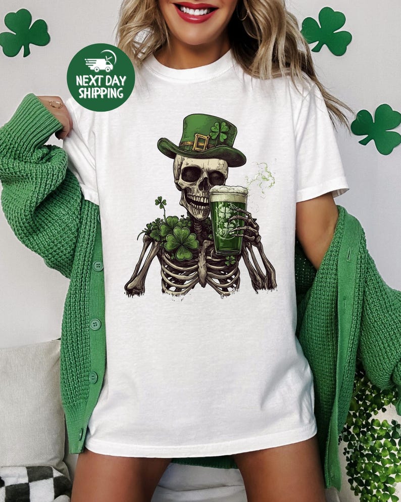 Puede incluir: Camiseta blanca con un esqueleto verde que lleva un sombrero de duende verde y sostiene una jarra de cerveza verde. El esqueleto est&aacute; rodeado de tr&eacute;boles verdes.