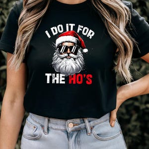 May include: Black short-sleeved t-shirt with a white graphic of Santa Claus wearing sunglasses and a red hat. The text "I do it for the ho's" is printed on the shirt.