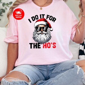 May include: Pink t-shirt featuring a graphic of Santa Claus wearing sunglasses and the text "I do it for the ho's".