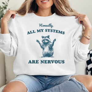 Puede incluir: Sudadera blanca con un gr&aacute;fico de mapache y el texto "Honestamente, todos mis sistemas est&aacute;n nerviosos".