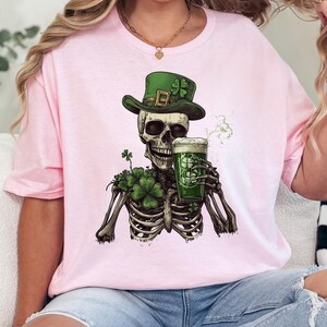 Puede incluir: Camiseta rosa con un esqueleto verde que lleva un sombrero de duende verde y sostiene una jarra de cerveza verde. El esqueleto est&aacute; rodeado de tr&eacute;boles verdes.