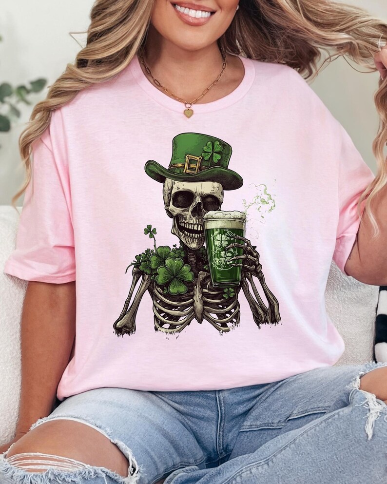 Puede incluir: Camiseta rosa con un esqueleto verde que lleva un sombrero de duende verde y sostiene una jarra de cerveza verde. El esqueleto est&aacute; rodeado de tr&eacute;boles verdes.