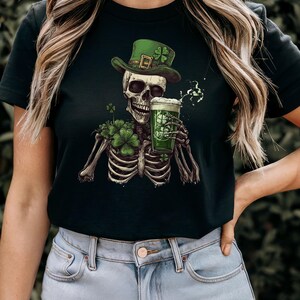 Puede incluir: Camiseta negra con un esqueleto verde que lleva un sombrero de duende verde y sostiene una jarra de cerveza verde. El esqueleto est&aacute; rodeado de tr&eacute;boles verdes.