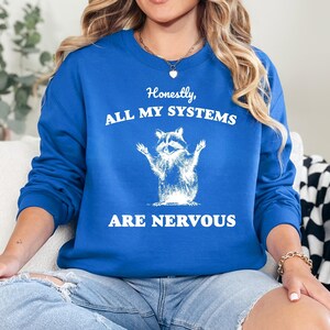 Puede incluir: Una sudadera azul real con un gr&aacute;fico blanco de un mapache con el texto "Honestamente, todos mis sistemas est&aacute;n nerviosos".