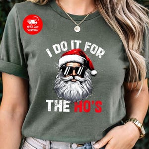 May include: Green t-shirt featuring a white graphic of Santa Claus wearing sunglasses and a white beard. The text "I do it for the ho's" is printed below the image.