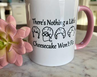 Puede incluir: Taza de cerámica blanca con interior y asa rosas. La taza presenta el texto "There's Nothing a Little Cheesecake Won't Fix" en negro, junto con dibujos de porciones de tarta de queso. Una suculenta rosa está en primer plano.