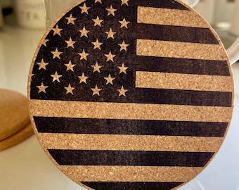 Puede incluir: Posavasos redondo de corcho con un diseño de bandera estadounidense grabado con láser. La bandera presenta franjas negras y un campo de estrellas negro. Una taza blanca está en el fondo. El texto "Goto LaserImaginations.com" está en la parte inferior.