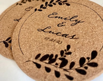 May include: Two round cork coasters with a laser-etched design. The coasters feature the names "Emily & Lucas" and the date "07.21.23" within a circular border, adorned with a botanical leaf pattern. The coasters are light brown.