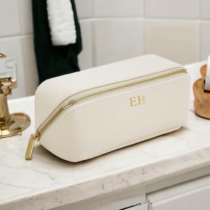 Può includere: Un beauty case bianco con cerniera, accessori dorati e le iniziali "EB" in rilievo sul davanti.