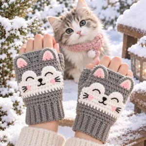 May include: A pair of gray fingerless gloves featuring a crocheted cat face design with white accents, pink inner ears, and black embroidered whiskers. The gloves have a ribbed cuff and a smiling cat expression on the back of each hand.