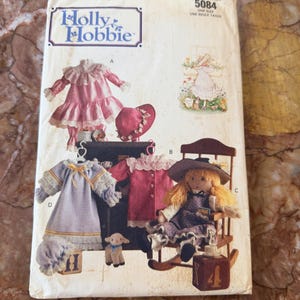 May include: A Butterick Holly Hobbie sewing pattern envelope 5084 featuring illustrations of doll clothing including a pink ruffled dress, a purple dress with lace trim, a red coat, and a doll sitting in a wooden rocking chair with a toy lamb.