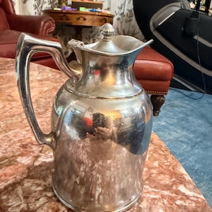 May include: A silver-colored pitcher with a handle and a lid. The pitcher has a bulbous body and a curved spout. The handle is large and arched. The pitcher is sitting on a marble-like surface. The pitcher is likely used for serving beverages.
