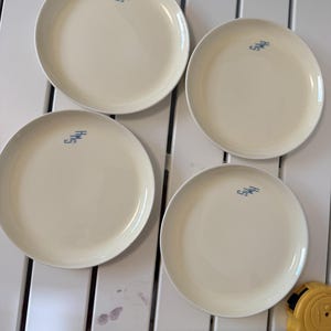 May include: Four round, cream-colored plates, each featuring the blue text "HMS" printed near the rim. A yellow measuring tape is positioned at the bottom, indicating a diameter of approximately 9 inches for the plates.