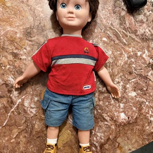 May include: A doll with brown hair and blue eyes wearing a red short-sleeved shirt with a blue and gray horizontal stripe, a small car graphic, and a Little Tikes logo. The doll wears blue denim cargo shorts, white socks, and tan boots.