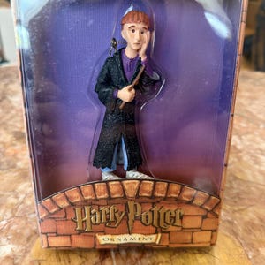 May include: A Ron Weasley figurine ornament inside a clear plastic display box. The character has red hair, a purple shirt, and a black textured robe, holding a wand. The box features a brick-patterned base with the text Harry Potter ORNAMENT.