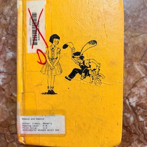 May include: A vintage yellow book titled "Beezus and Ramona" with black illustrations of children. The cover features a girl in a dress and a child with bunny ears. The book has a library sticker with author and reading level information.