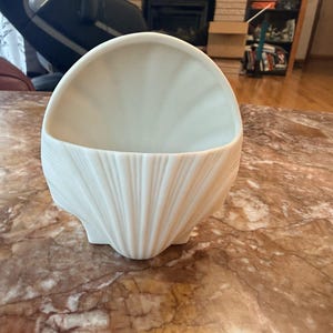 May include: A matte off-white ceramic wall pocket shaped like a large seashell. The vessel features a semi-circular opening at the top and a textured exterior with vertical ridges radiating from the base, mimicking the natural form of a scallop shell.