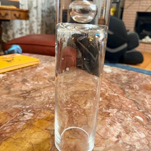 May include: A clear glass bottle with a spherical orb suspended inside. The bottle has a cylindrical base and a flared top, with a decorative element above the orb. The glass is transparent, allowing a view of the interior.