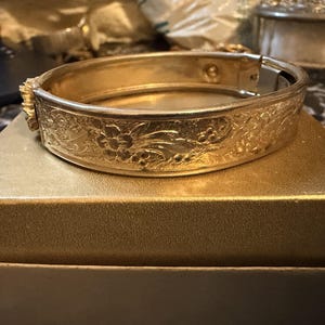 May include: A gold-toned metal hinged bangle bracelet featuring an intricate embossed floral pattern across the outer surface. The bracelet has a polished finish and a secure clasp mechanism visible on the side, showcasing a detailed relief design.