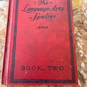 May include: A vintage red book titled "The Language Arts Speller" with black lettering and a black border. The book is labeled "Book Two" and has the author's name, McCall, printed below the title. The book's cover shows signs of wear.
