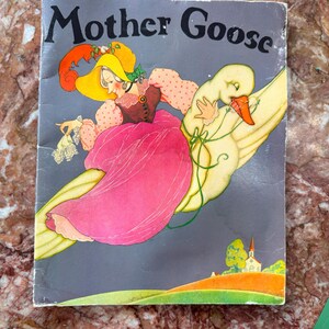 May include: Vintage "Mother Goose" book cover featuring a whimsical illustration. A woman in a pink dress and yellow hat rides a goose against a gray background. The title is in black, with a colorful landscape at the bottom.