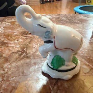 May include: A white ceramic elephant figurine with a raised trunk and painted details. The elephant has black, red, and green accents. The figurine is on a white base.